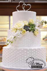 Whipped cream tops this beautiful cake, the top tier smooth, the bottom two tiers decorated with swiss dots. Fresh flowers and succulents adorn this cake as well as a cake topper with two hearts.