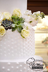Whipped cream tops this beautiful cake, the top tier smooth, the bottom two tiers decorated with swiss dots. Fresh flowers and succulents adorn this cake as well as a cake topper with two hearts.
